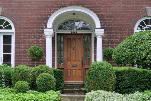 Portico entrance of traditional brick house surrounded by shrubbery, with elegant wood grain front door with sidelights