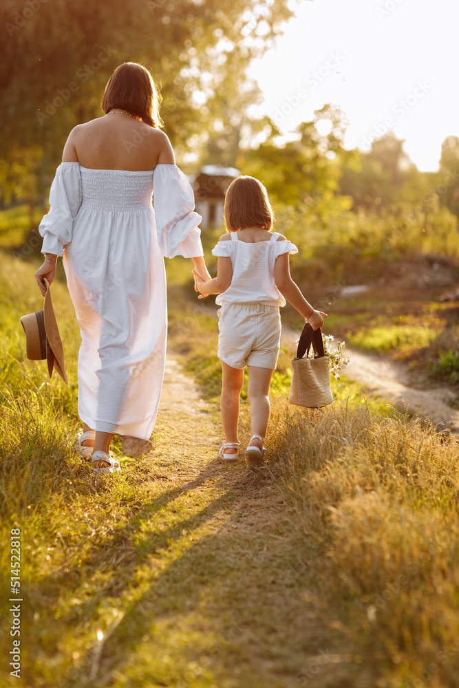 Fototapeta premium young mother is holding her child's daughter hand and walking in green meadow on a summer sunny day. Family having fun together. Childhood, parenting, happiness concept. mother's day. back view