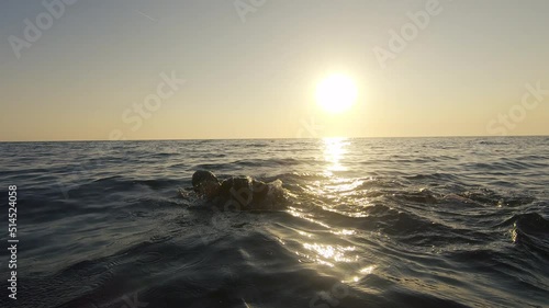 Male swimmer training in open sea at sunset, wide tracking shot. Sportsman exercising in choppy sea and sun shining on horizon. Athlete workout in ocean