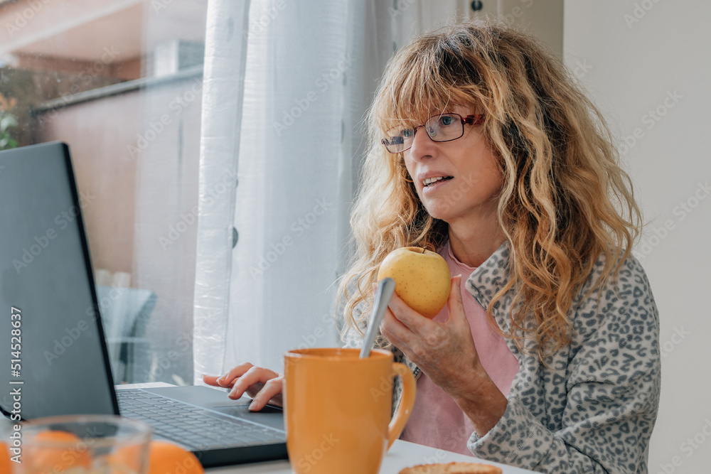 middle aged woman having breakfast at home with laptop middle aged woman having breakfast at home with laptop