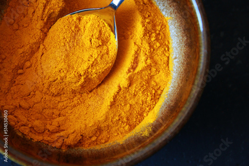close-up of yellow turmeric spice powder with a spoon in a brown ceramic bowl on dark background, top view of turmeric powder as cooking spice