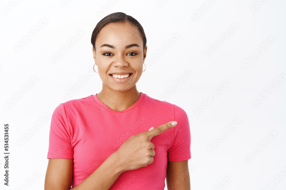 Close up of enthusiastic latina woman smiles, points finger right, shows advertisement, stands in pink tshirt over white background Close up of enthusiastic latina woman smiles, points finger right, shows advertisement, stands in pink tshirt over white background