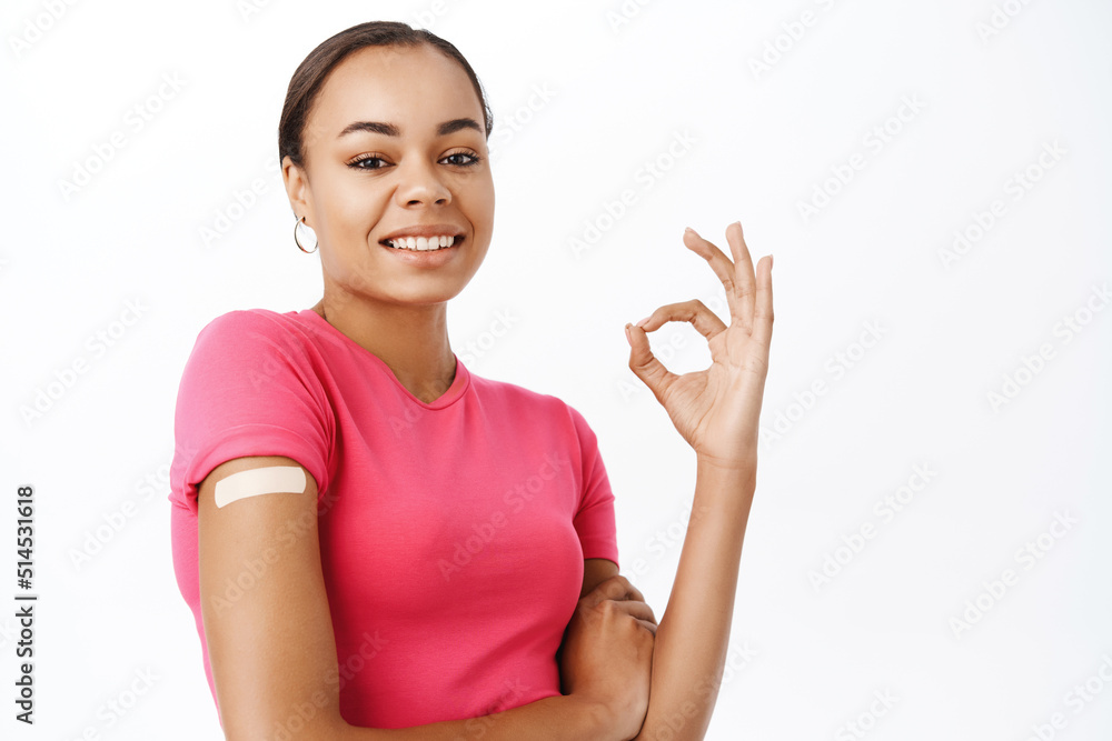 Portrait of healthy smiling woman shows okay sign after covid 19 vaccination, has patch on shoulder after coronavirus vaccine shot, stands over white background Portrait of healthy smiling woman shows okay sign after covid 19 vaccination, has patch on shoulder after coronavirus vaccine shot, stands over white background