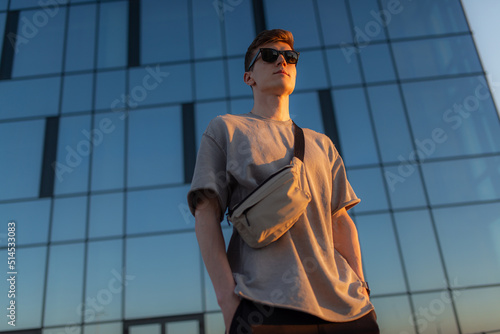 Portrait of handsome tourist hipster man in sunglasses with bag handy, standing guy near the building in downtown. Calm relaxing moment, summer vacation concept