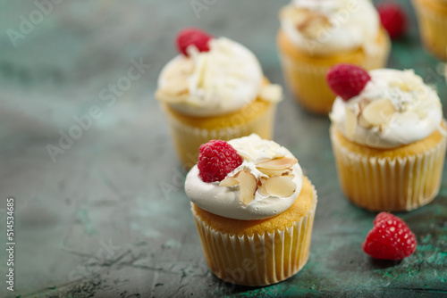 On a dark green background, there are many appetizing cupcakes with airy white cream, decorated with raspberries and almonds. Sweet food, treat for holiday, birthday.