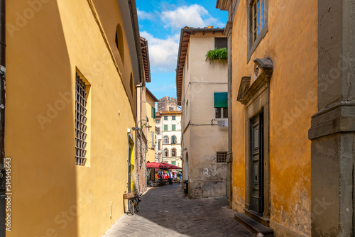 A narrow street of homes, shops and cafes inside the walled medieval town of Lucca, Italy, with the Tower of Basilica San Frediano in view behind.