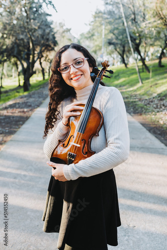 Smiling brunette woman with glasses holding and hugging a violin outside in a park road. Vertical