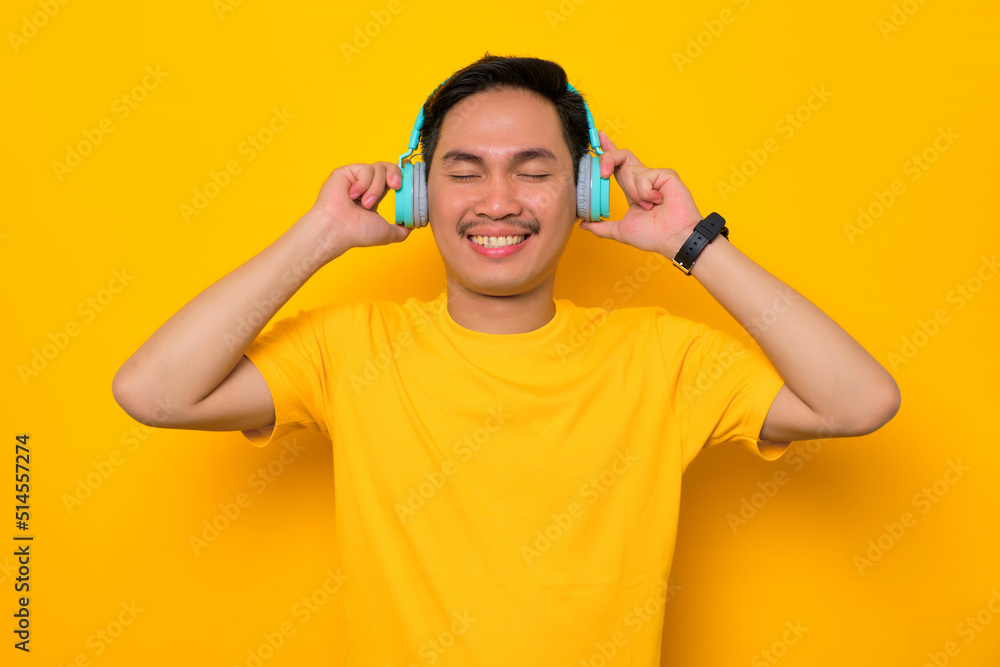 Relaxed young Asian man in casual t-shirt listening music in headphones isolated on yellow background. People emotions lifestyle concept Relaxed young Asian man in casual t-shirt listening music in headphones isolated on yellow background. People emotions lifestyle concept