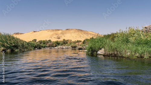 The riverbed bends between banks overgrown with green vegetation. An orange sand dune against a clear blue sky. Reflection on calm water. Egypt. Nile. Aswan © Вера 