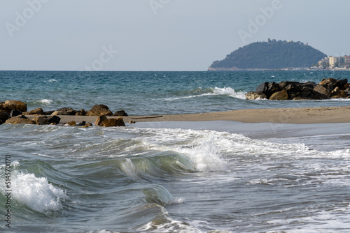 Beach on the Italian Riviera with sun, sea and stones in the sea water. Rough Sea and Foamy Waves on Shore at Mediterranean Coast in Sunrise