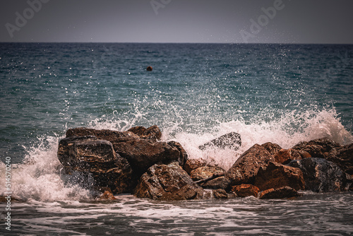 Beach on the Italian Riviera with sun, sea and stones in the sea water. Rough Sea and Foamy Waves on Shore at Mediterranean Coast in Sunrise