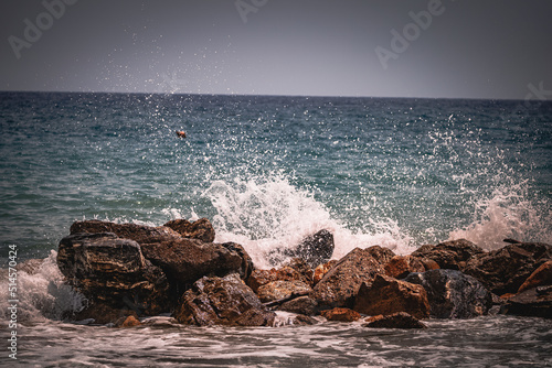 Beach on the Italian Riviera with sun, sea and stones in the sea water. Rough Sea and Foamy Waves on Shore at Mediterranean Coast in Sunrise