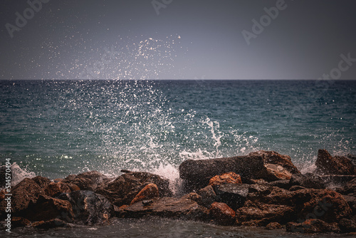 Beach on the Italian Riviera with sun, sea and stones in the sea water. Rough Sea and Foamy Waves on Shore at Mediterranean Coast in Sunrise