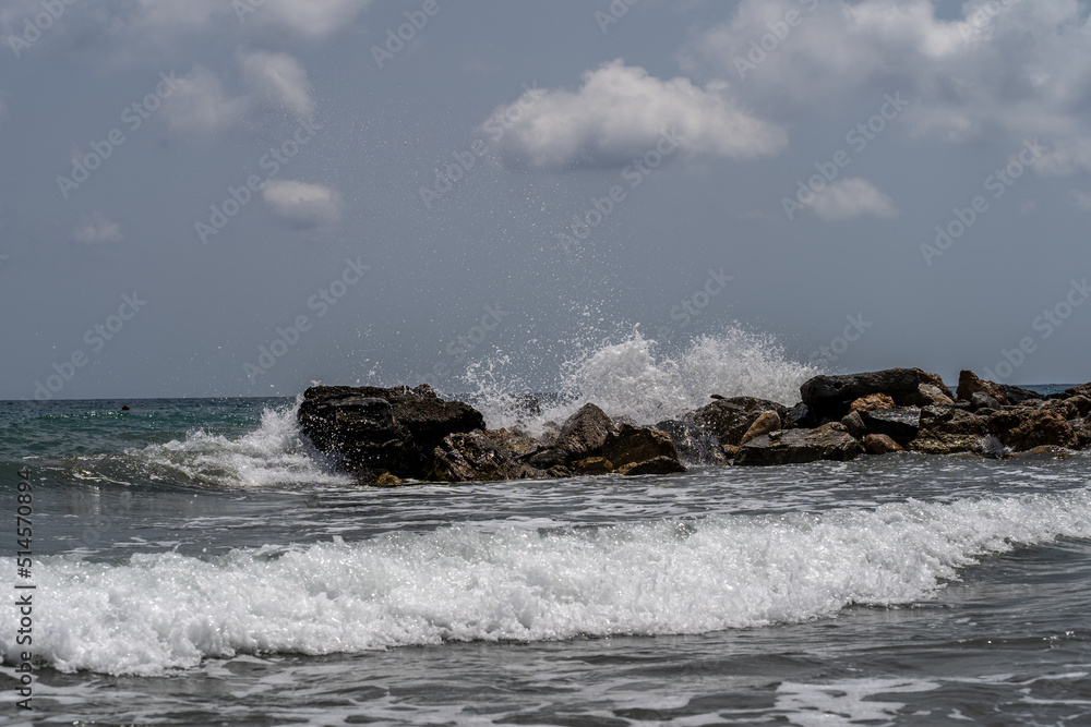 Obraz premium Beach on the Italian Riviera with sun, sea and stones in the sea water. Rough Sea and Foamy Waves on Shore at Mediterranean Coast in Sunrise