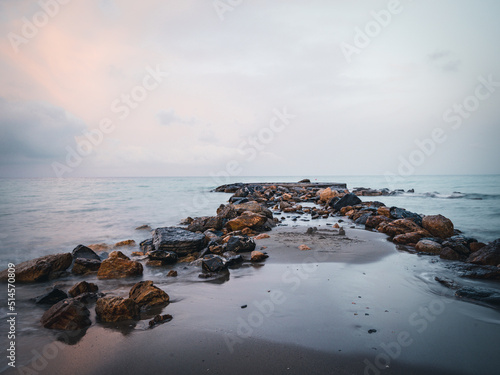 Beach on the Italian Riviera with sun, sea and stones in the sea water. Rough Sea and Foamy Waves on Shore at Mediterranean Coast in Sunrise