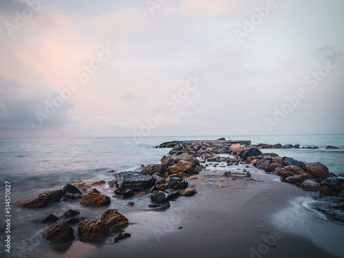 Beach on the Italian Riviera with sun, sea and stones in the sea water. Rough Sea and Foamy Waves on Shore at Mediterranean Coast in Sunrise