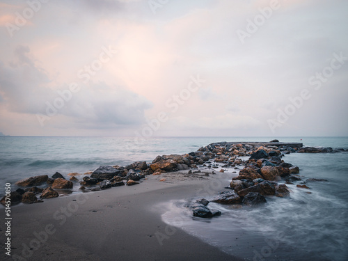 Beach on the Italian Riviera with sun, sea and stones in the sea water. Rough Sea and Foamy Waves on Shore at Mediterranean Coast in Sunrise