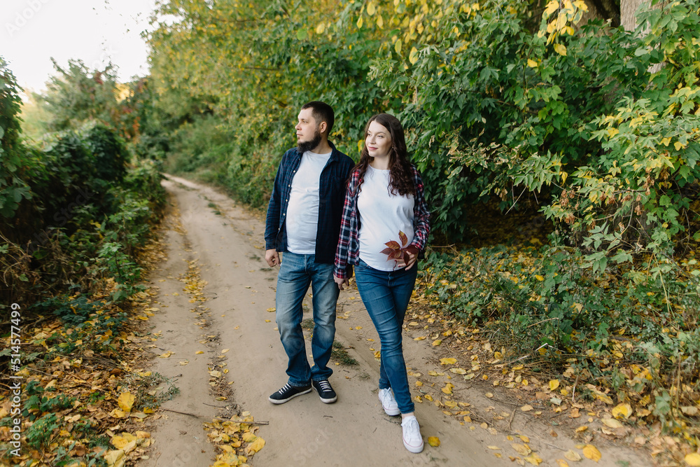 Obraz premium Beautiful young couple of future parents stand surrounded by autumn nature in the countryside outside the city and look into the distance. Pregnant woman holds an autumn leaf near her stomach. High
