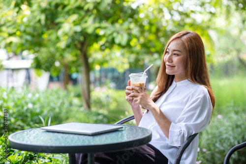 Portrait image a young asian woman holding and drinking iced coffee with laptop on the table in the outdoors