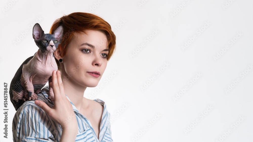 Portrait of caucasian redhead young woman with Sphynx Cat sitting on her shoulder. Female hipster in striped white and blue shirt. Studio shot on white background, copy space on right. Part of series.