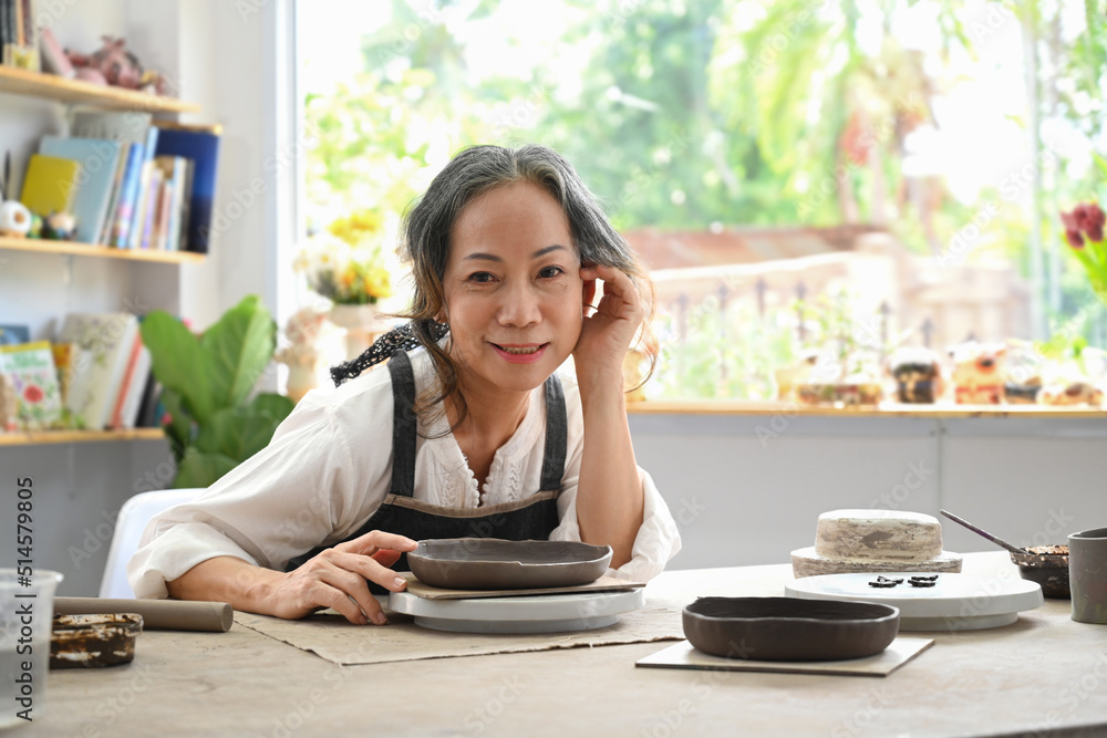 Smiling mature woman with handicraft crockery sitting in pottery workshop Smiling mature woman with handicraft crockery sitting in pottery workshop