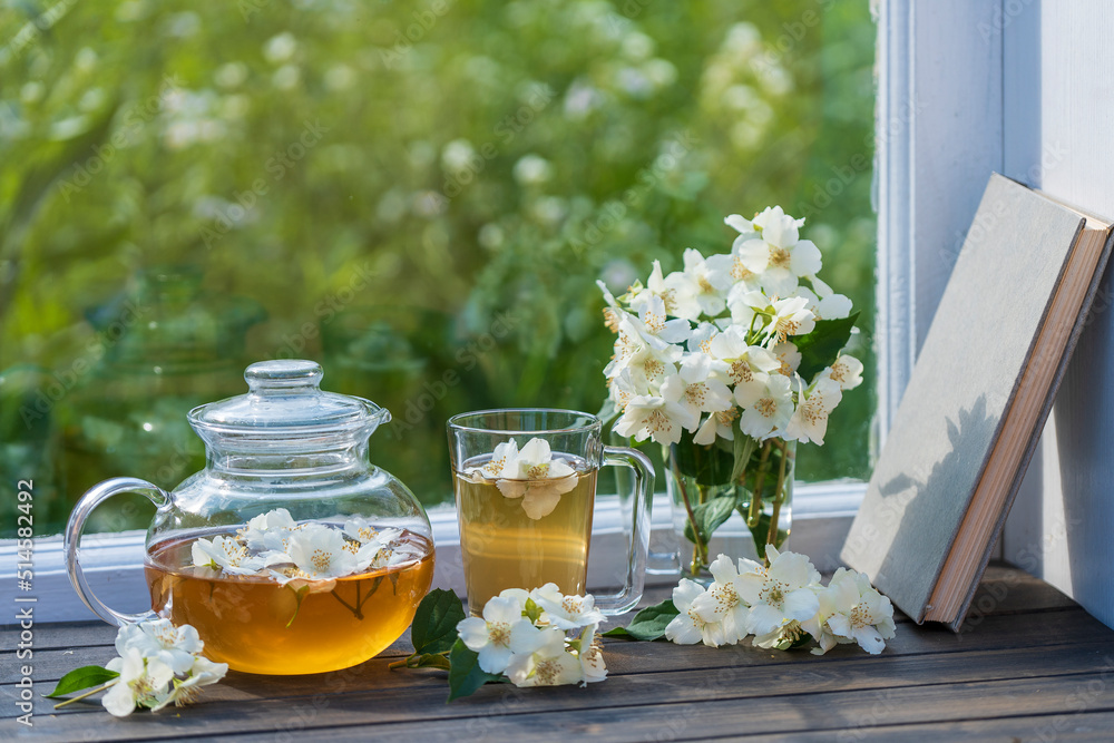 Hot herbal tea in glass teapot, cup and beautiful bouquet of jasmine flowers on windowsill at summer day near garden Hot herbal tea in glass teapot, cup and beautiful bouquet of jasmine flowers on windowsill at summer day near garden