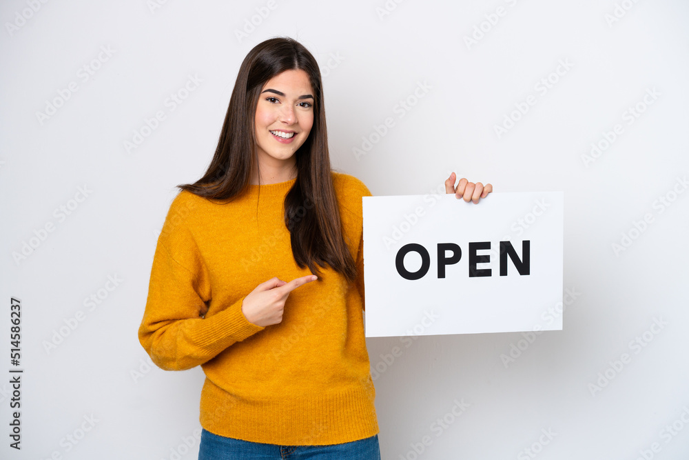 Young Brazilian woman isolated on white background holding a placard with text OPEN and pointing it Young Brazilian woman isolated on white background holding a placard with text OPEN and pointing it
