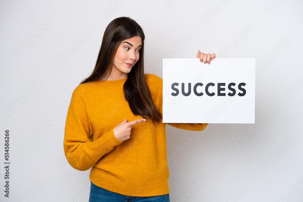Young Brazilian woman isolated on white background holding a placard with text SUCCESS and pointing it Young Brazilian woman isolated on white background holding a placard with text SUCCESS and pointing it
