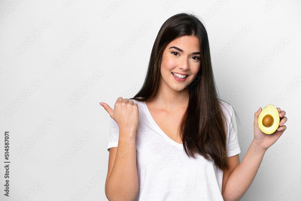 Young Brazilian woman holding an avocado isolated on white background pointing to the side to present a product Young Brazilian woman holding an avocado isolated on white background pointing to the side to present a product