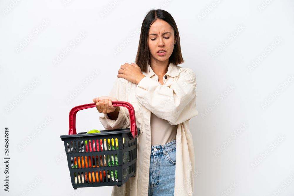Young woman holding a shopping basket full of food isolated on white background suffering from pain in shoulder for having made an effort Young woman holding a shopping basket full of food isolated on white background suffering from pain in shoulder for having made an effort