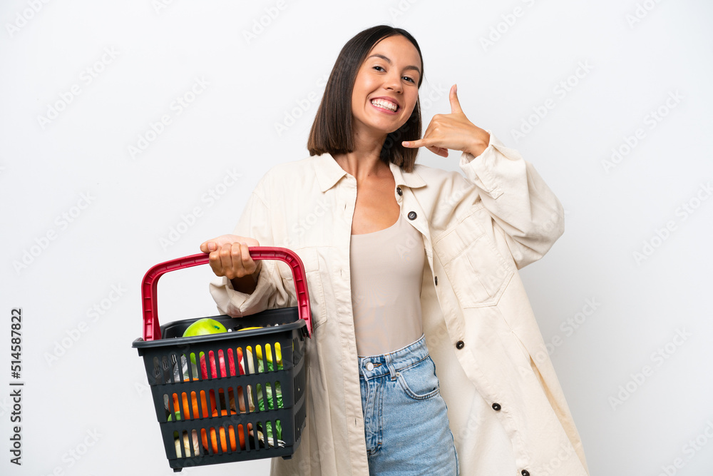 Young woman holding a shopping basket full of food isolated on white background making phone gesture. Call me back sign Young woman holding a shopping basket full of food isolated on white background making phone gesture. Call me back sign