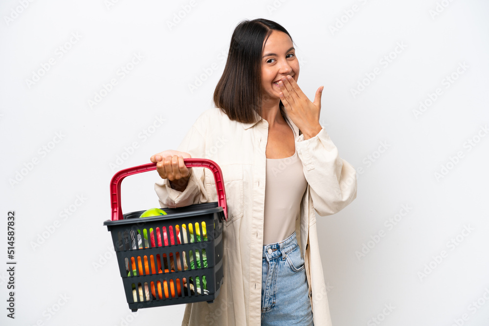 Young woman holding a shopping basket full of food isolated on white background happy and smiling covering mouth with hand Young woman holding a shopping basket full of food isolated on white background happy and smiling covering mouth with hand