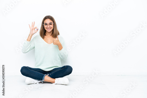 Young caucasian woman sitting on the floor isolated on white background showing ok sign and thumb up gesture