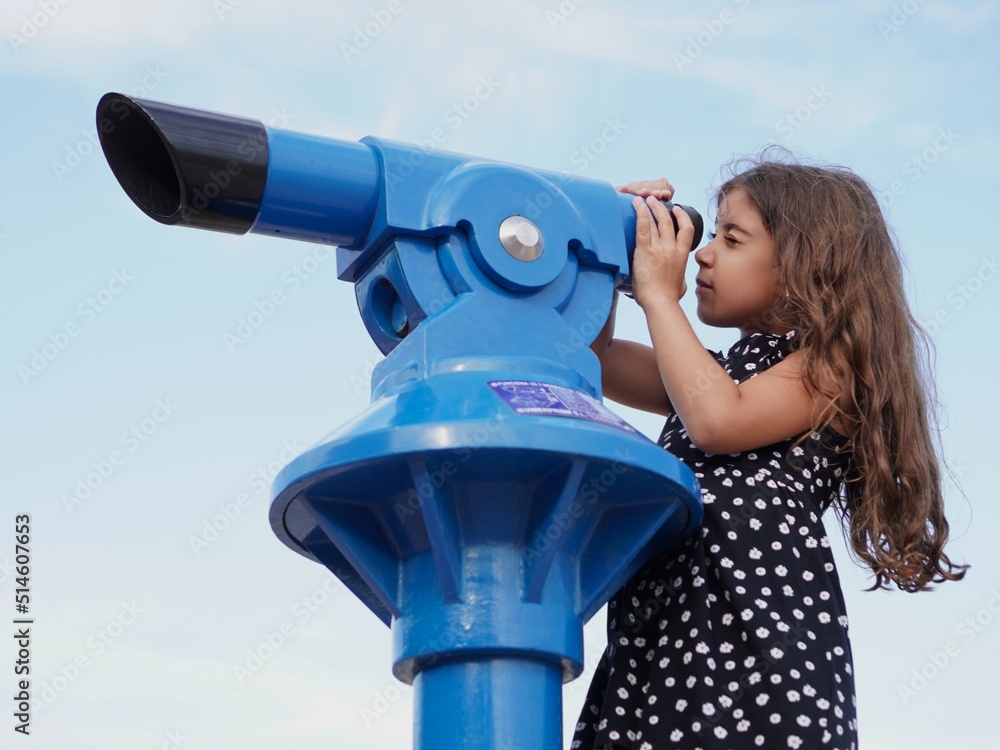 Caucasian girl looks curiously through a blue telescope at the surrounding landscape with a cloudless blue sky background, adventurous and curious girl concept