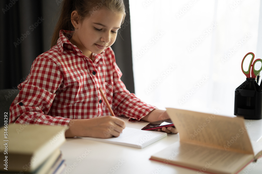 Close up focused little girl using phone studying online, sitting at table at home, serious child holding smartphone, watching webinar, writing notes, homeschooling Close up focused little girl using phone studying online, sitting at table at home, serious child holding smartphone, watching webinar, writing notes, homeschooling