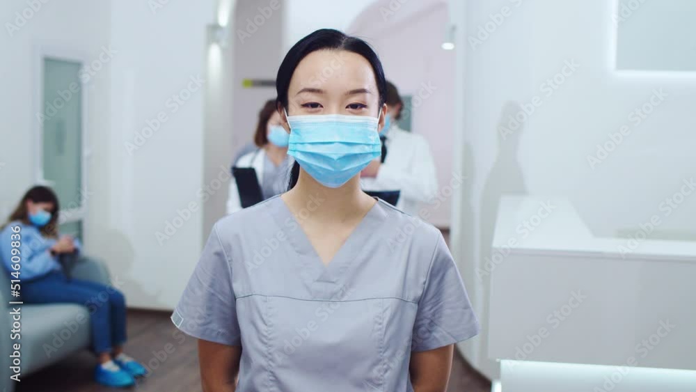 Portrait of Asian happy young beautiful female hospital practitioner wearing protective mask while standing in private clinic hall. Healthcare worker, microbiology, pharmaceutical, research, Portrait of Asian happy young beautiful female hospital practitioner wearing protective mask while standing in private clinic hall. Healthcare worker, microbiology, pharmaceutical, research,