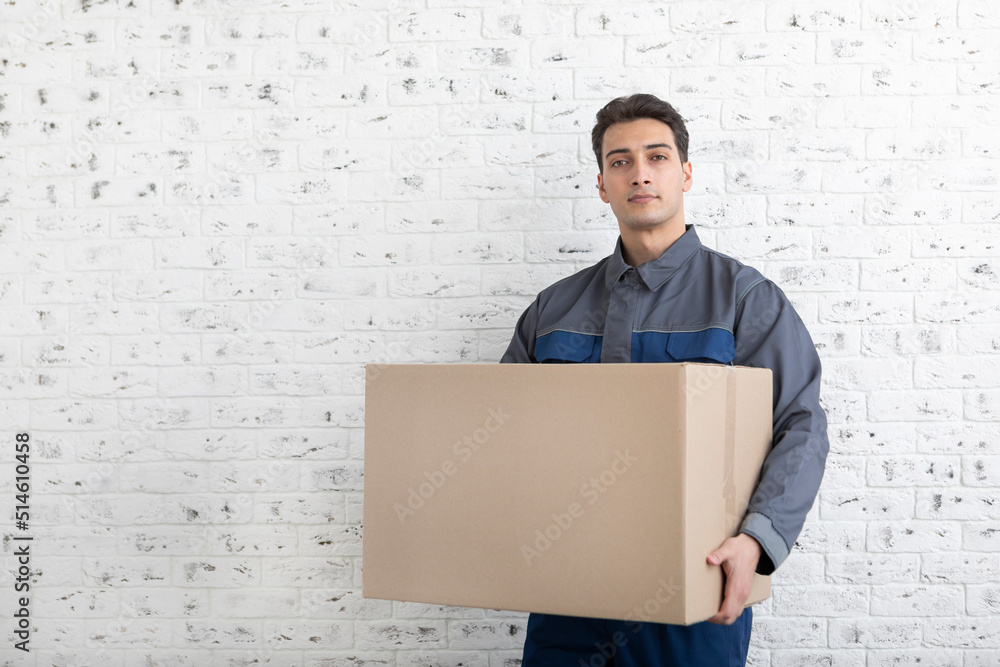Confident male from moving service holding box beside white brick wall background Confident male from moving service holding box beside white brick wall background