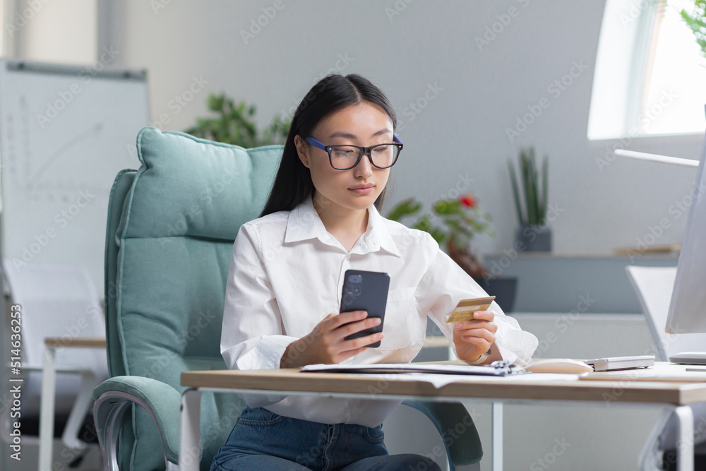 Beautiful and confident Asian business woman working in the office, making banking transactions, using the phone and banking application, holding a credit card, for online shopping. Beautiful and confident Asian business woman working in the office, making banking transactions, using the phone and banking application, holding a credit card, for online shopping.