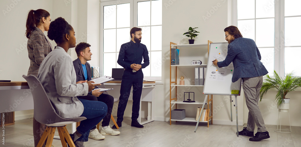 Business coach giving presentation by office whiteboard. Smart experienced man meets with team of people, shares his expertise and teaches to use analysis and problem solving methods