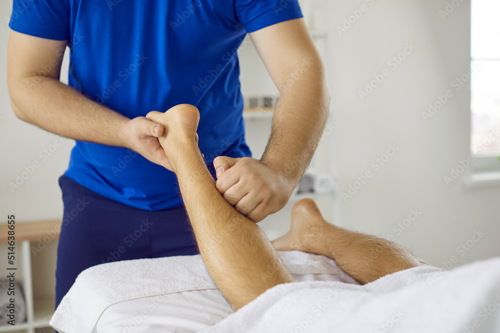 Masseur massaging patient's leg. Professional physiotherapist, osteopath, massagist, or chiropractor massaging ankle, shin and calf muscles of young man. Cropped shot. Physiotherapy treatment concept