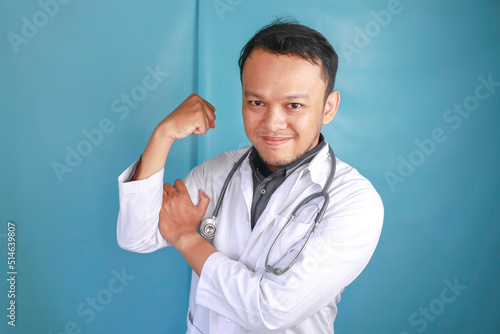 A portrait of a young Asian male doctor showing his biceps isolated by a blue background