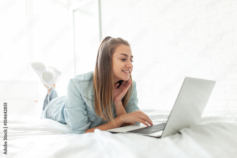Young smiling woman using laptop while lying on bed in room on bright day Young smiling woman using laptop while lying on bed in room on bright day