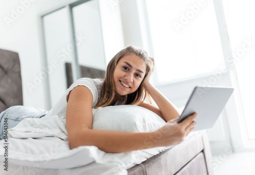 Young beautiful caucasian woman uses a tablet while lying on a bed in a bright room