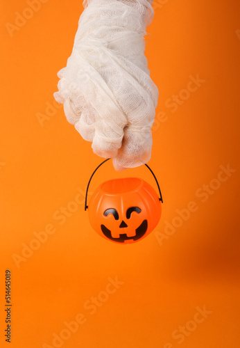 Trick or treat. Happy Halloween. Mummy's hand in white intes holds bucket for candies on orange background photo