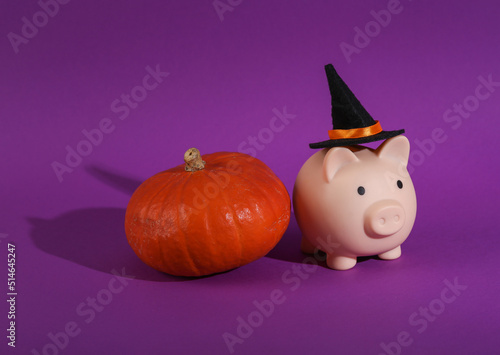 Halloween still life. Piggy bank in witch hat and pumpkin on purple background