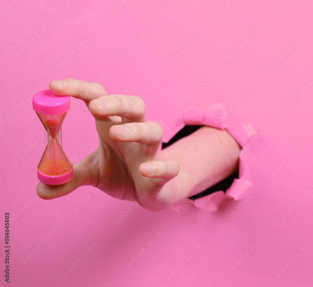 Female hand holds hourglass through torn hole pink paper. Concept art. Female hand holds hourglass through torn hole pink paper. Concept art.