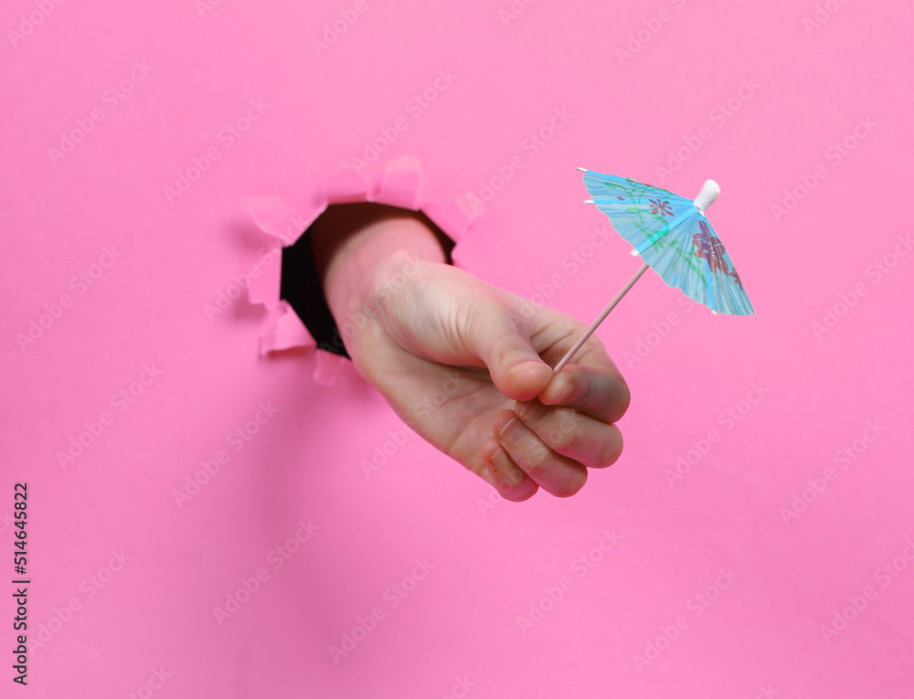 Female hand holds cocktail umbrella through torn hole pink paper. Concept art. Minimalism.