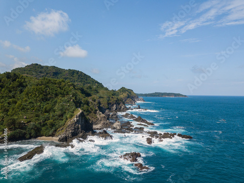 Cliffs overgrown by trees that are directly adjacent to the sea. There are rocks that are washed away by the waves with blue sky. The sea water looks blue. Pengilon Hill, Yogyakarta, Indonesia