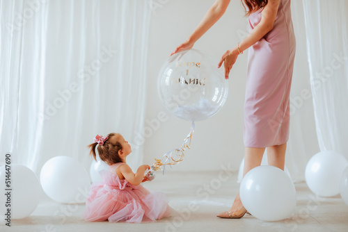 Cute one year girl in pink beautiful dress and slim mother posing in the studio with big balloon. Focus is at the girl. Party concept