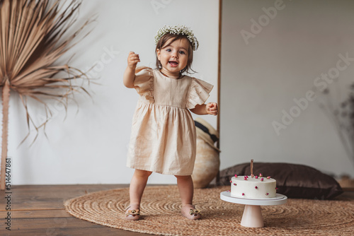 Obraz na płótnie Cute one year girl in beige dress and cute flower crown posing with white birthday cake in the studio
